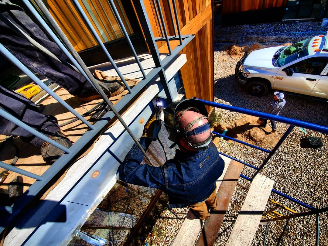 Overhead view of a welder TIG welding a steel balcony railing against a wooden building